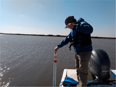 A man on a boat checks a water gauge that stands as a white pole protruding from the water