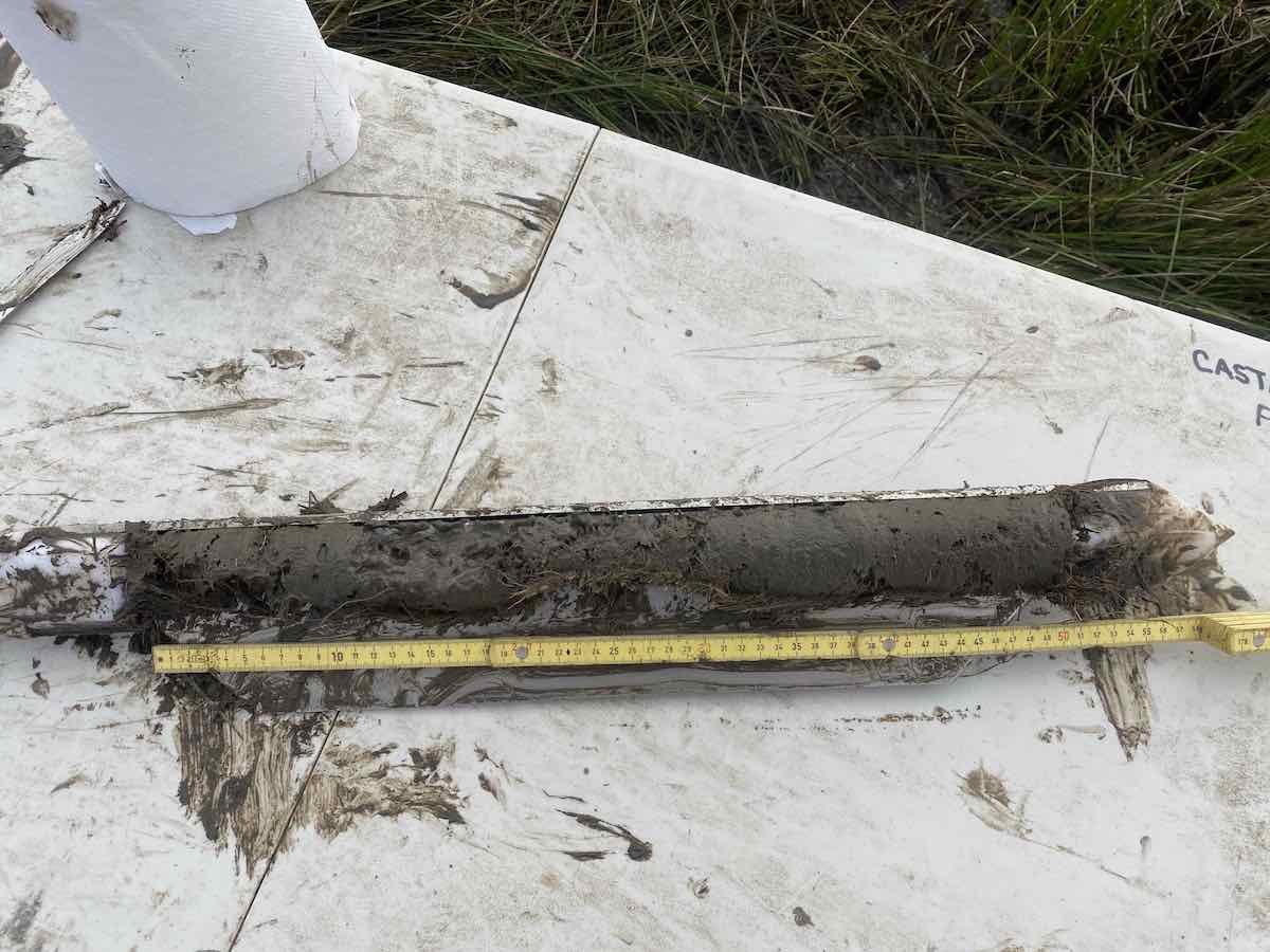 A white pipe encased in soil sits on the ground with a tape measuring the length of the soil