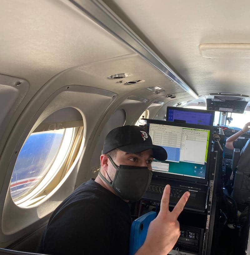 Luis sits in front of a monitor in the aircraft cabin and makes the victory sign with his hand
