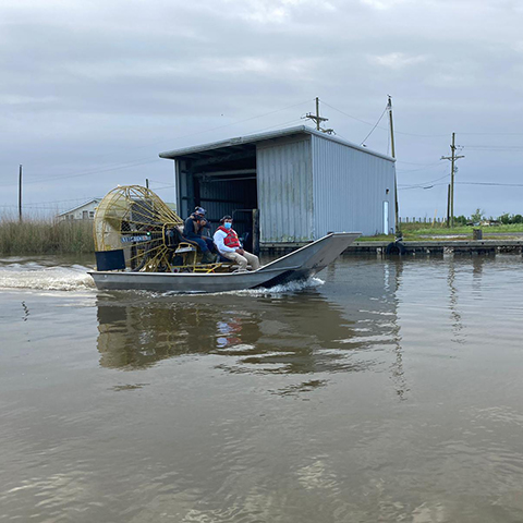 Two people sit in an airboat on the water, with a shed in the background