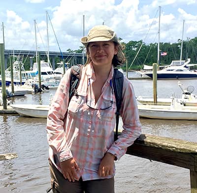 Elena Solohin stands in front of a dock on a sunny day with boats behind her