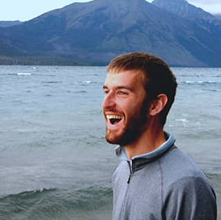 John smiles with mouth open looking into the distance, with water and a mountain behind him